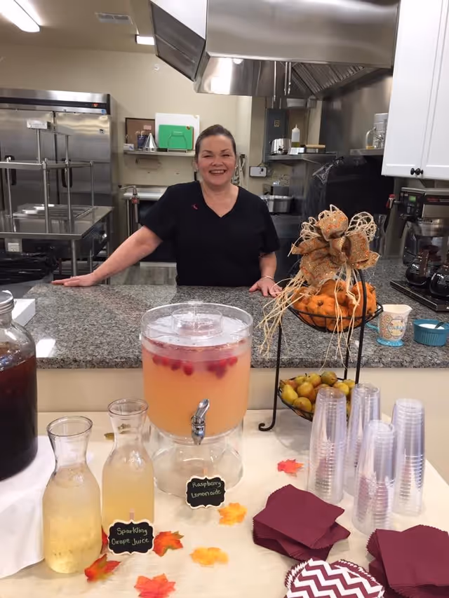 A woman in a black shirt stands behind a granite counter in a kitchen area, smiling. In front of her on the counter are a large dispenser of raspberry lemonade, two glass carafes labeled sparkling grape juice and raspberry lemonade, stacks of clear plastic cups, maroon napkins, and a two-tiered fruit basket with oranges and pears. The kitchen has stainless steel appliances and equipment in the background.