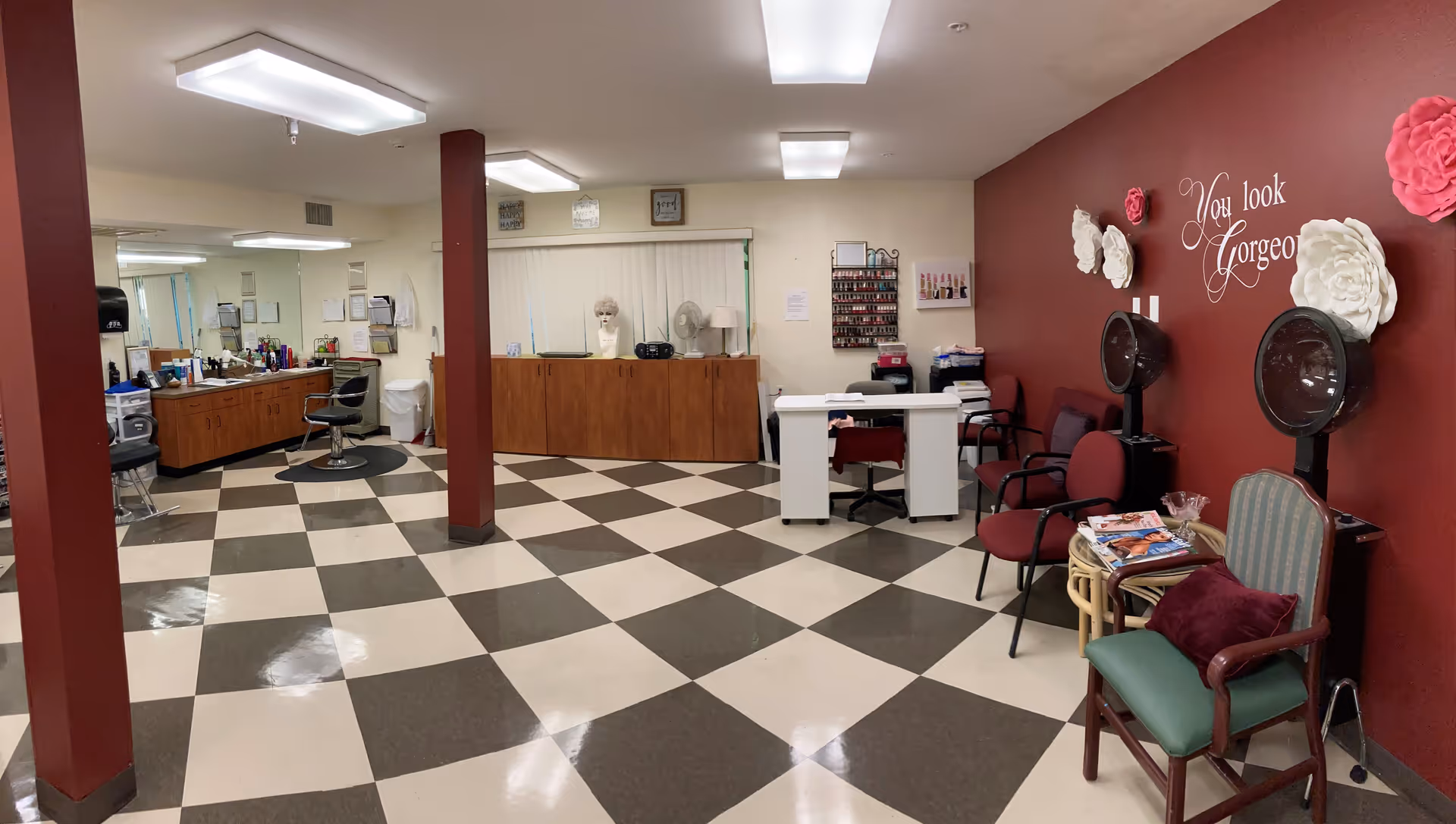 Interior view of a beauty salon area in a senior living facility with checkered floor tiles, maroon and white walls, salon chairs, hair dryers, a manicure table, nail polish racks, and decorative flowers on the wall with the phrase 'You look Gorgeous'.