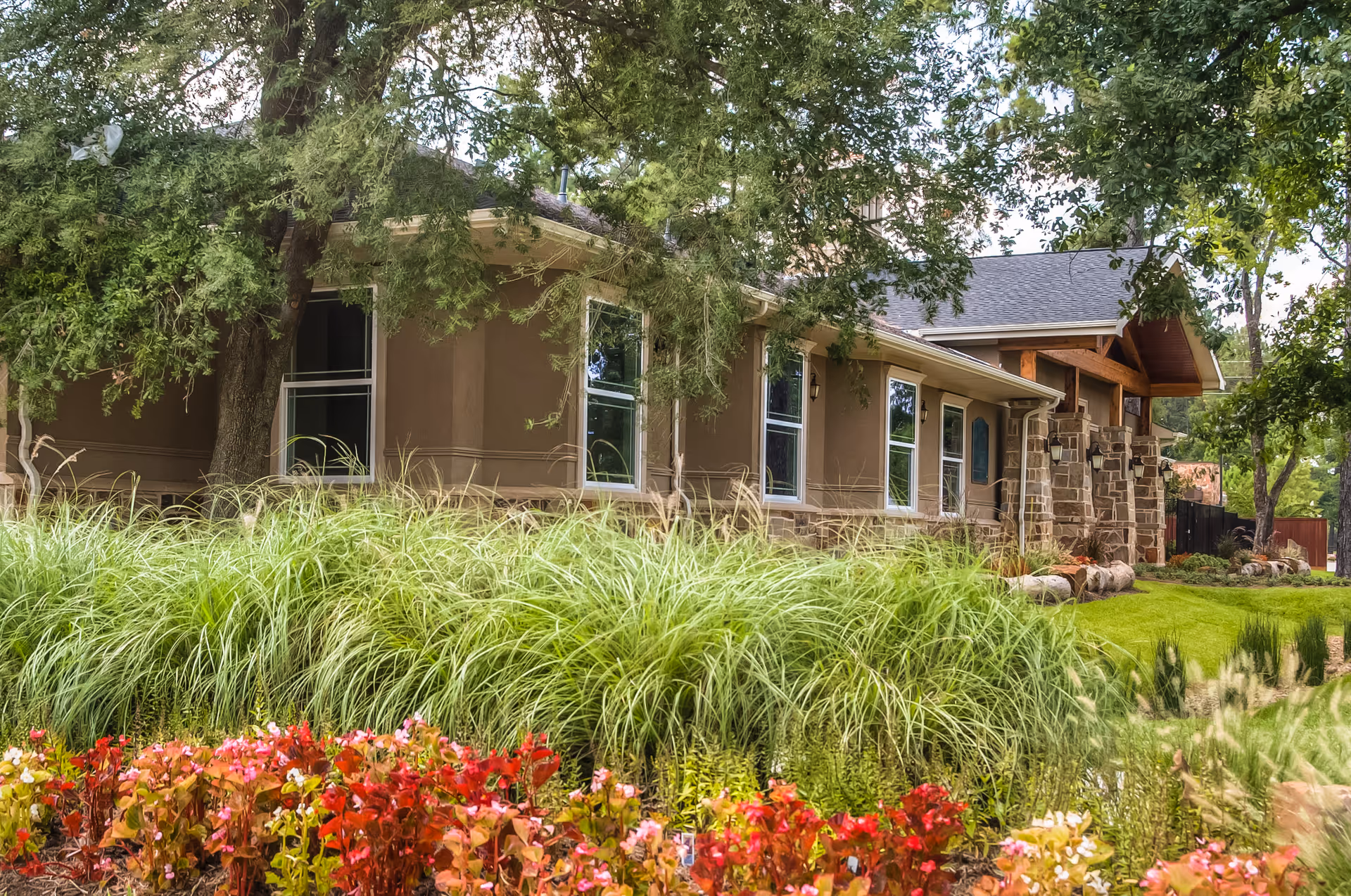 Exterior view of a single-story building with beige walls and stone accents, surrounded by lush greenery including tall grasses, colorful flowers, and large trees providing shade.