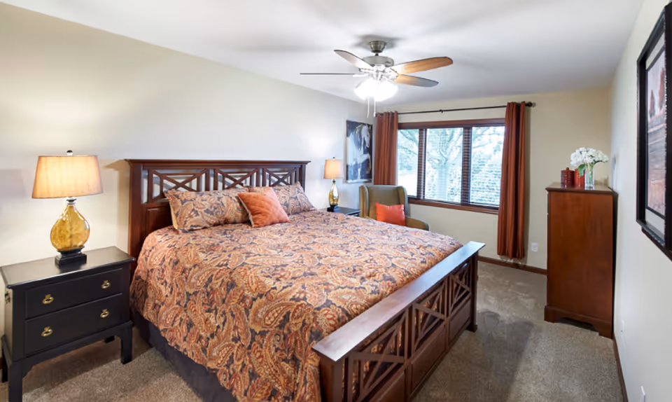 A cozy bedroom with a large wooden bed featuring a patterned bedspread and pillows. There are two bedside tables with lamps, a ceiling fan with lights, a window with brown curtains, a green armchair with an orange pillow, a wooden dresser with decorative items, and a framed picture on the wall.