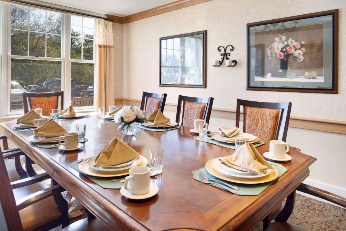 A dining room with a wooden table set for six people. Each place setting includes a plate, a folded beige napkin, a glass, a coffee cup with a saucer, and silverware. The room has large windows with beige curtains, framed artwork on the walls, and a small floral centerpiece on the table.