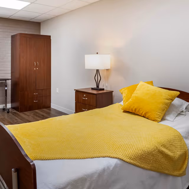 A simple nursing home bedroom with a single bed covered by a yellow throw, a bedside table and lamp, and a wooden wardrobe.