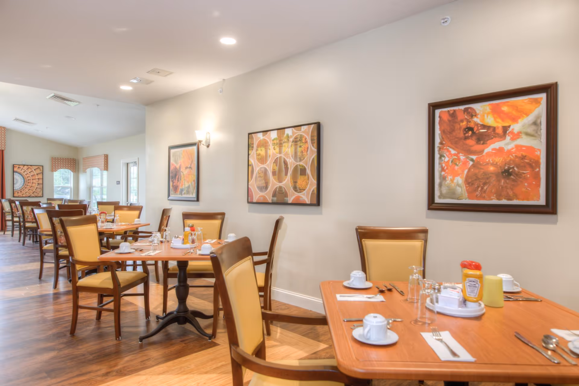 A dining room in a senior living facility with wooden tables and chairs featuring yellow cushions. Tables are set with white cups, saucers, utensils, and condiments including ketchup and mustard. The room has light-colored walls adorned with framed abstract artwork and large windows with valances allowing natural light to enter.