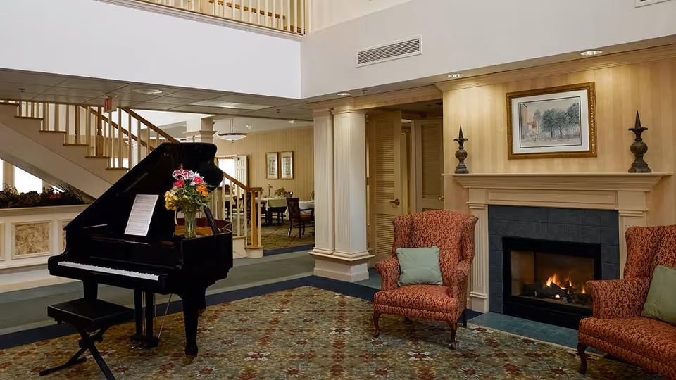 A cozy living room area in a senior living facility featuring a black grand piano with a vase of flowers and sheet music on it, two red patterned armchairs with green cushions, a lit fireplace with a framed painting above it, and a staircase leading to an upper level. In the background, there is a dining area with tables and chairs.
