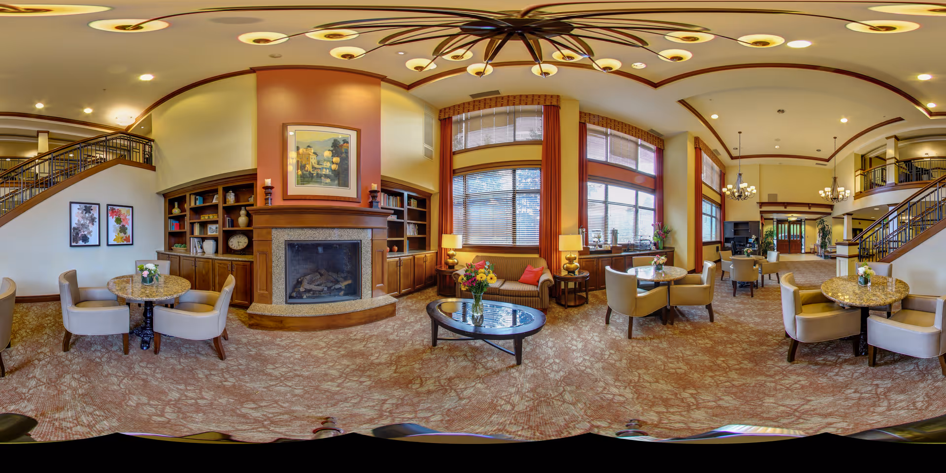 A spacious and well-lit senior living facility common area featuring a large stone fireplace with built-in wooden bookshelves on either side. The room has multiple seating arrangements including round tables with chairs and a sofa with side tables and lamps. Large windows with red curtains allow natural light to fill the room. The ceiling has decorative lighting fixtures and the carpet has a patterned design. A staircase with wooden railings is visible on the left and right sides of the room.
