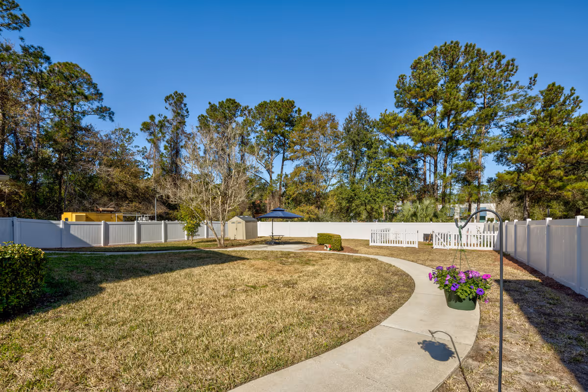 A fenced outdoor garden area with a curved concrete pathway, a hanging flower pot with purple flowers, a small table with an umbrella, and trees in the background under a clear blue sky.