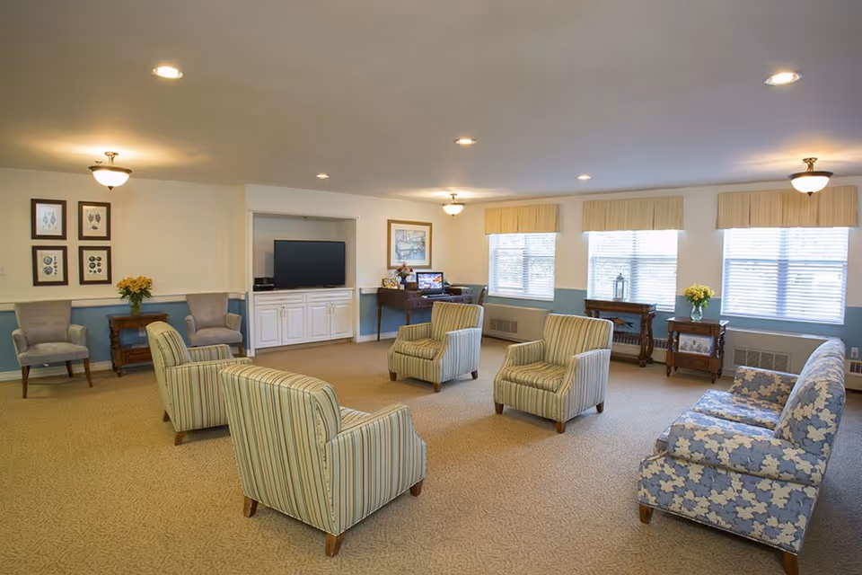 Spacious senior living lounge with several upholstered armchairs and sofas arranged around a TV and windows.