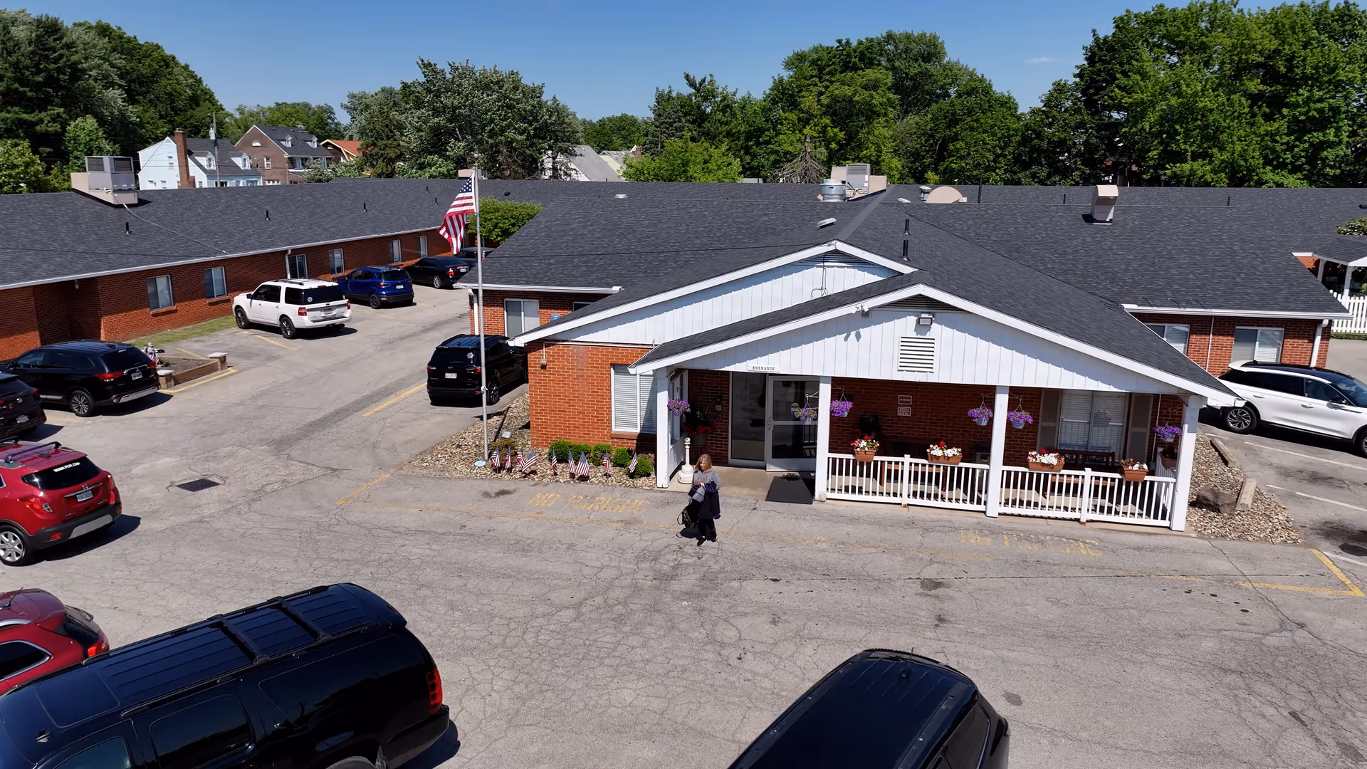 Exterior view of a single-story brick building with a covered porch decorated with hanging flower baskets and potted plants. Several cars are parked in the surrounding parking lot, and an American flag is flying on a flagpole near the entrance. A person is walking in front of the building. Trees and other houses are visible in the background under a clear blue sky.