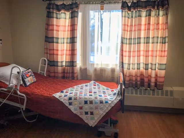 A single bed with a red quilt and a colorful patchwork blanket folded at the foot. The bed has a white pillow and a decorative pillow with a floral design. Behind the bed is a window with sheer white curtains and striped curtains in shades of red, pink, and green. The room has wooden flooring and a beige wall.
