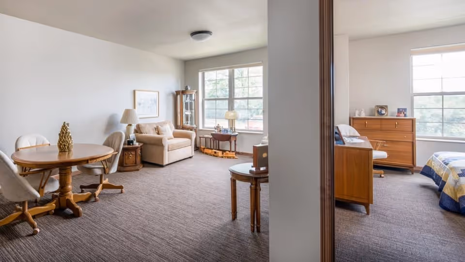 Bright senior living apartment interior showing a sitting area with sofa and table, a round dining table, and a partially visible bedroom separated by a wall.