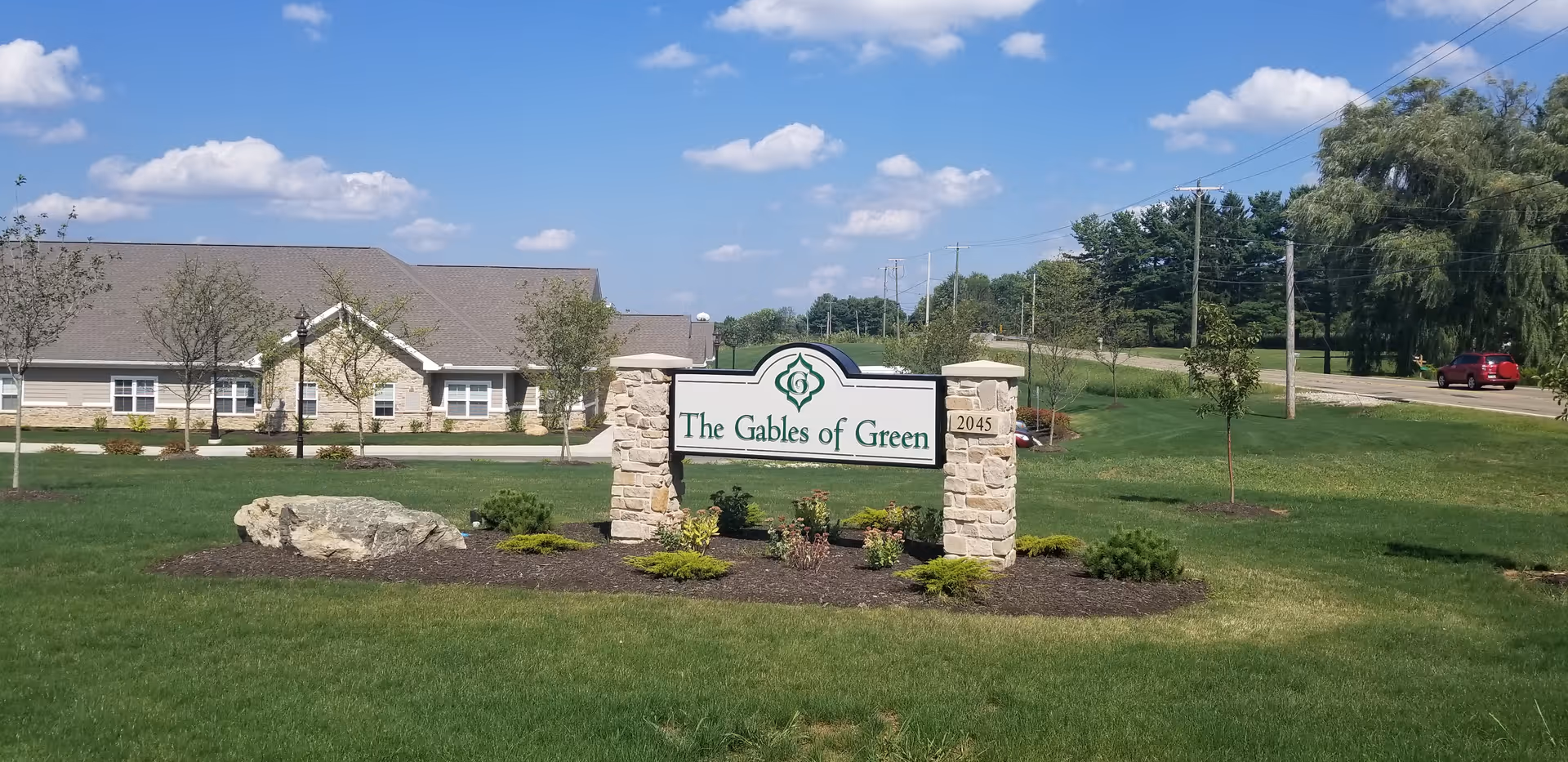 A landscaped lawn with a stone and wood sign that reads 'The Gables of Green' in front of a single-story building under a blue sky with scattered clouds. There are small trees and shrubs around the sign and a road with a red car in the background.
