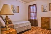 Furnished bedroom with a twin bed, bedside lamp on a wooden nightstand, a wooden dresser, wall art and hardwood floors.