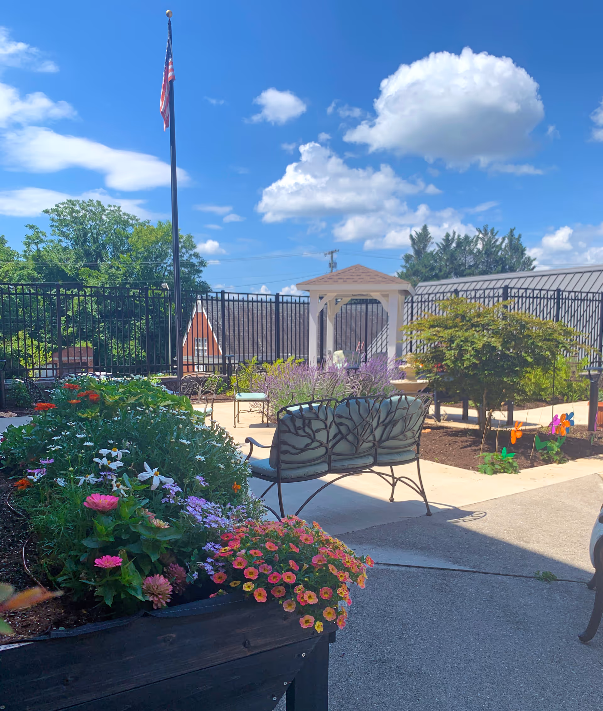 Outdoor garden area with colorful flowers in raised beds, metal benches with decorative backs, a small white gazebo, and an American flag on a tall flagpole under a blue sky with scattered clouds.