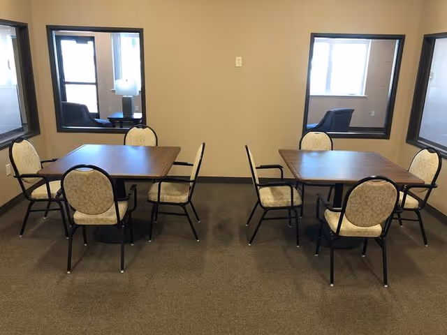 Two rectangular tables with upholstered chairs in a small communal dining room with interior windows.