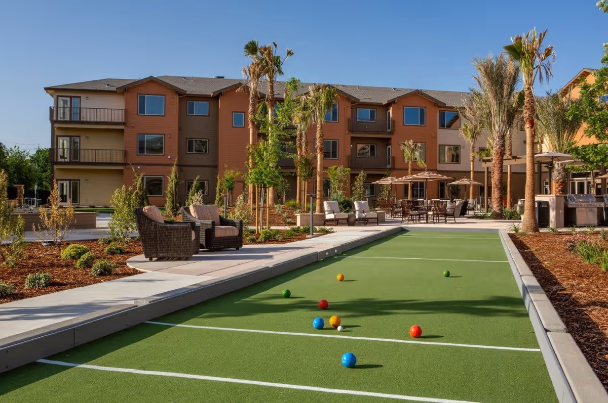 Outdoor bocce ball court with colorful balls scattered on the green playing surface, surrounded by patio seating with chairs and tables under umbrellas, palm trees, and a three-story residential building in the background under a clear blue sky.