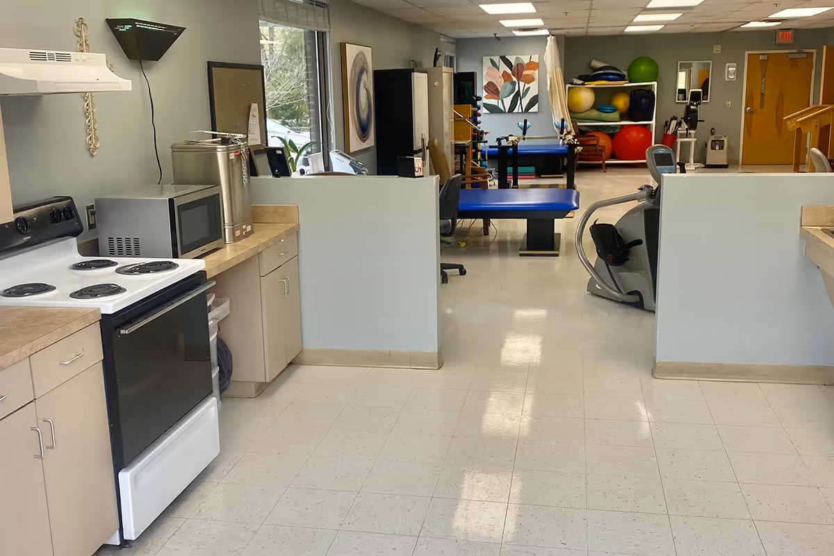Interior view of a facility room showing a kitchen area on the left with a stove, microwave, and countertop. Beyond the kitchen, there is an open space with exercise and therapy equipment including a stationary bike, therapy tables, and shelves with exercise balls. The room has tiled flooring, light-colored walls, and fluorescent ceiling lights.
