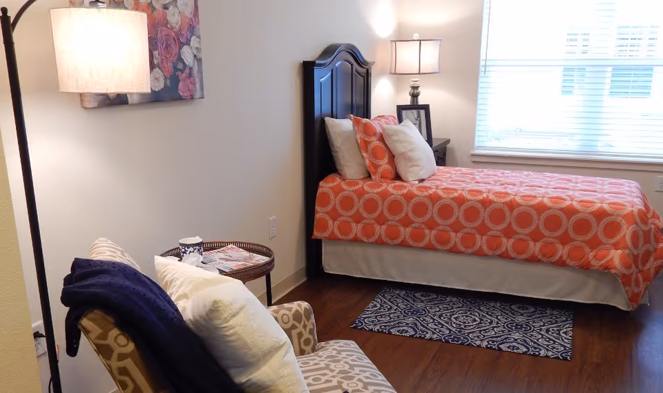 A cozy bedroom with a single bed covered in an orange bedspread with circular patterns. There are several pillows on the bed. Next to the bed is a nightstand with a lamp and a framed photo. A window with blinds allows natural light into the room. In the foreground, there is a patterned armchair with a dark blue throw blanket and white pillows. A small round side table holds a cup and some magazines. The floor is wooden with a small patterned rug near the bed.