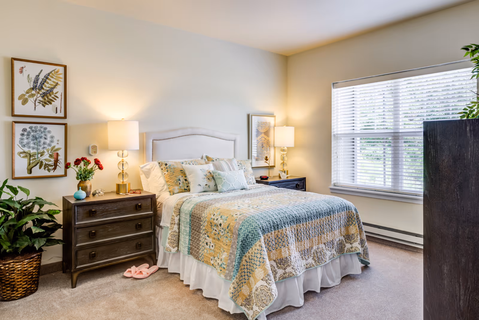 A cozy bedroom with a neatly made bed featuring a patchwork quilt in soft pastel colors. There are two bedside tables, each with a lamp and decorative items. A large window with white blinds lets in natural light. The room also has framed botanical artwork on the wall and a potted plant in a woven basket on the floor.