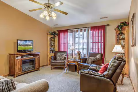A cozy living room with beige walls and carpeted floor, featuring a ceiling fan with lights, a flat-screen TV on a wooden stand with an electric fireplace, two brown leather armchairs, a beige sofa, a wooden side table with a lamp, and large windows with vertical blinds and red curtains.