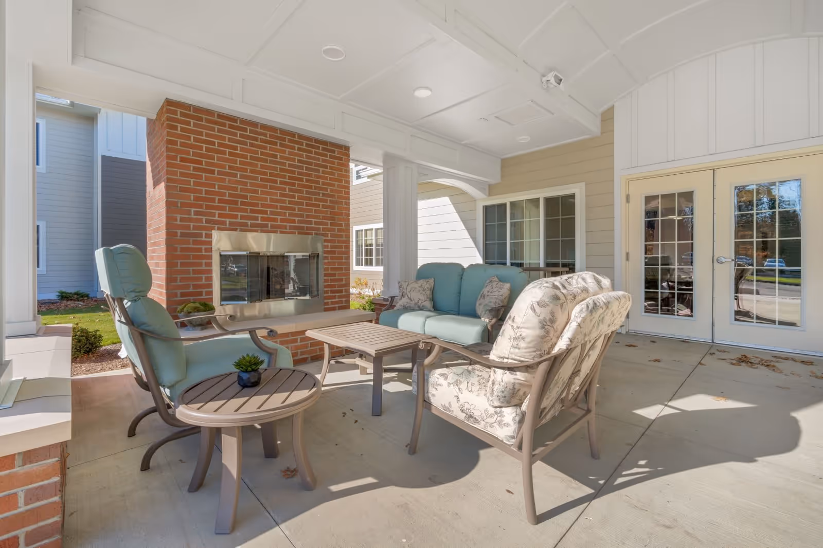 Covered outdoor seating area with cushioned chairs and a sofa arranged around a rectangular coffee table. There is a brick fireplace with a metal frame on one side and glass double doors leading inside the building. The area is well-lit with natural light and has a concrete floor.