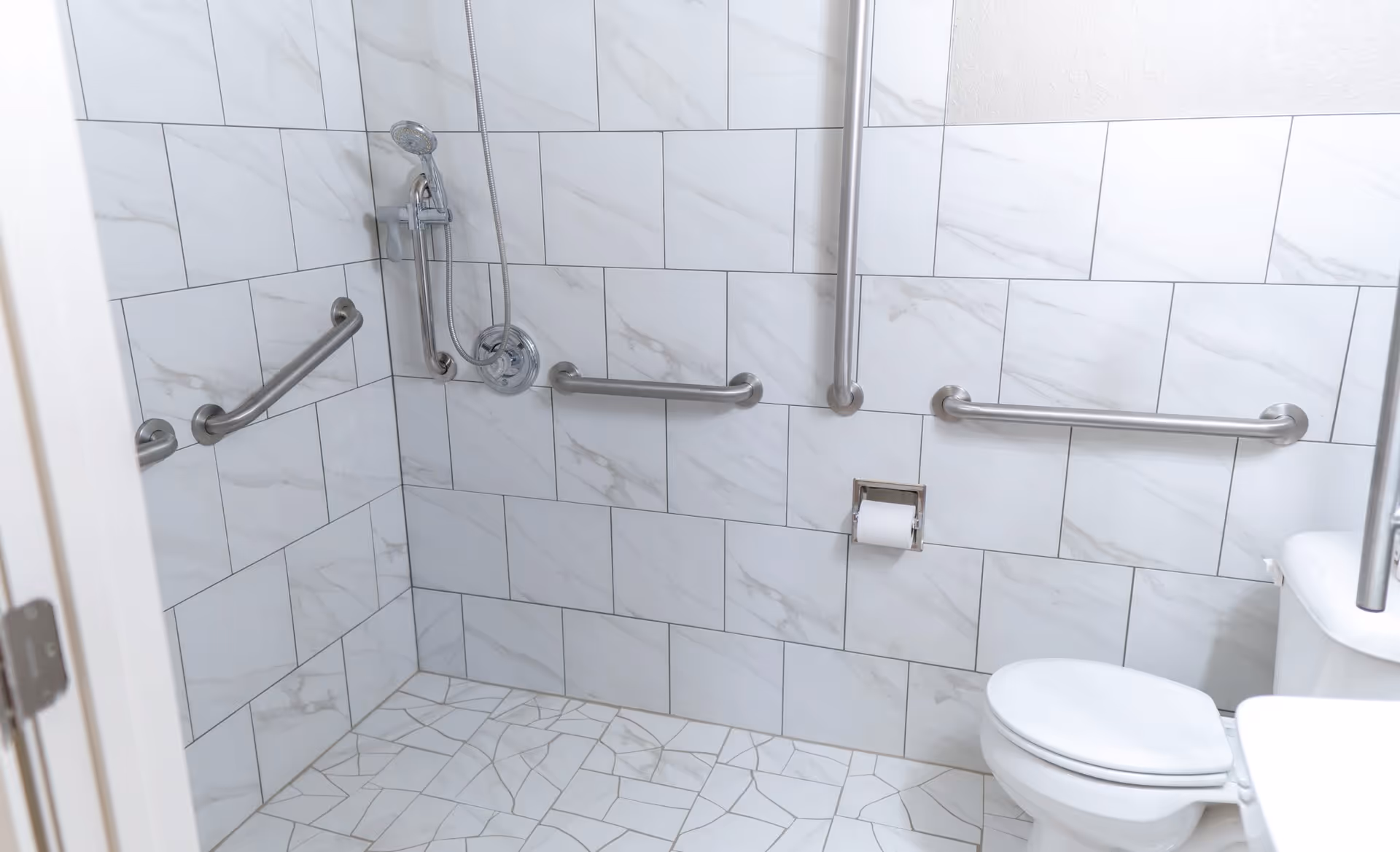 A bathroom with white marble-patterned tiles on the walls and floor, equipped with multiple stainless steel grab bars, a handheld showerhead, a toilet with a closed lid, and a toilet paper holder.