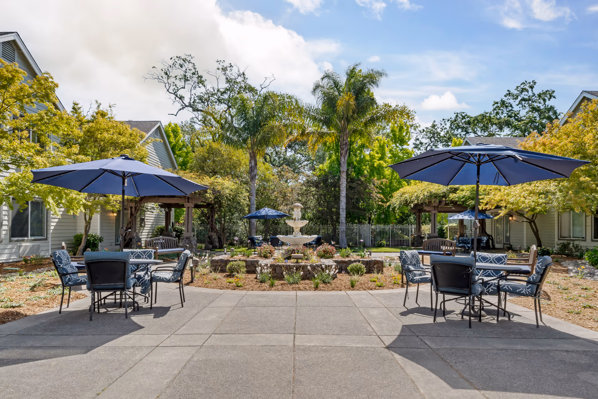 Outdoor patio area with two tables, each shaded by a large blue umbrella and surrounded by cushioned chairs. In the background, there is a stone fountain centered among landscaped plants and flowers, with palm trees and other greenery. Residential buildings with light-colored siding flank the patio on both sides under a partly cloudy blue sky.