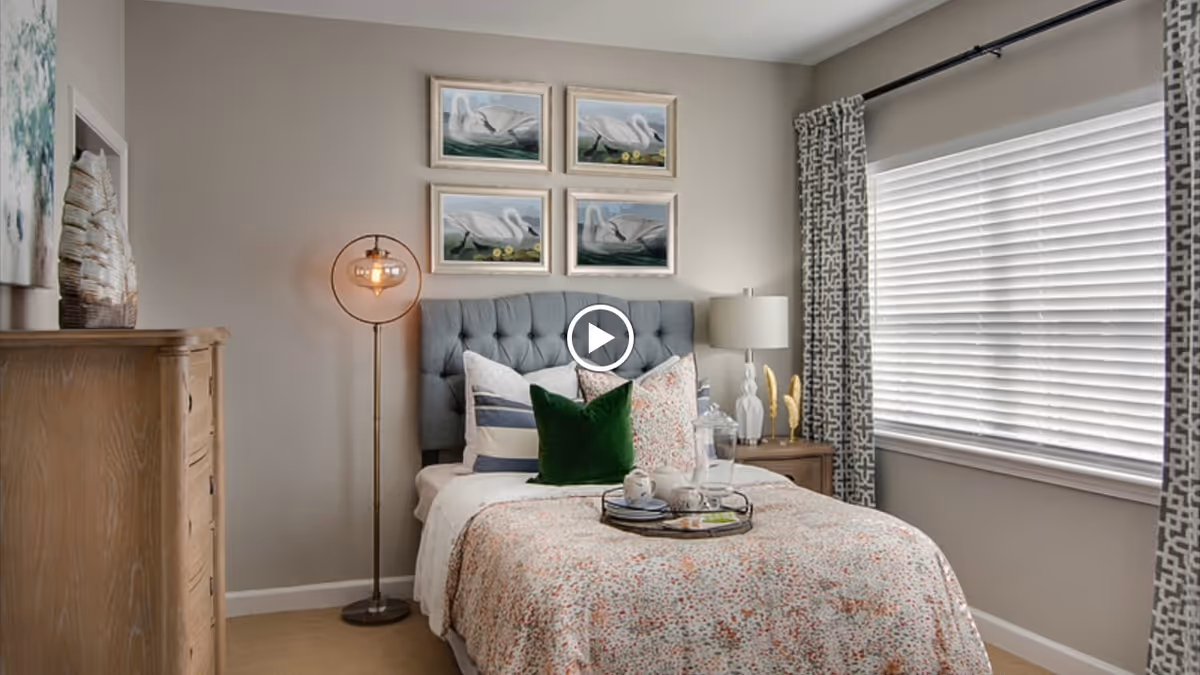 A cozy bedroom with a neatly made bed featuring a tufted gray headboard, multiple pillows including a green velvet one, and a floral-patterned bedspread. Above the bed are four framed pictures of swans. To the left of the bed is a wooden dresser and a modern floor lamp with an exposed bulb. To the right is a wooden nightstand with a white lamp and decorative items. A window with patterned curtains lets in natural light.