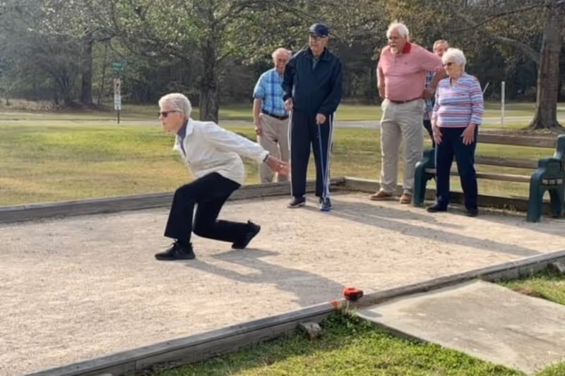 A group of elderly people outdoors playing bocce ball on a sandy court. One person is in the action of throwing a bocce ball while others watch. There are trees and grass in the background and a bench nearby.