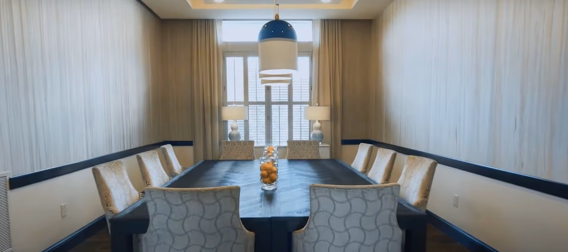 A modern dining room with a long rectangular dark wood table surrounded by ten upholstered chairs with patterned fabric. The room has light-colored walls with a subtle texture, two table lamps on a console table near a window with white shutters, and a large pendant light hanging above the center of the table. A glass vase filled with oranges is placed on the table.