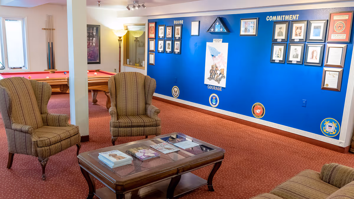 A cozy lounge area with two upholstered armchairs and a wooden coffee table with magazines on it. In the background, there is a blue wall decorated with framed photos, certificates, and military insignias under the words 'HONOR', 'COURAGE', and 'COMMITMENT'. A pool table with red felt is visible behind a white pillar, along with a floor lamp and a window.