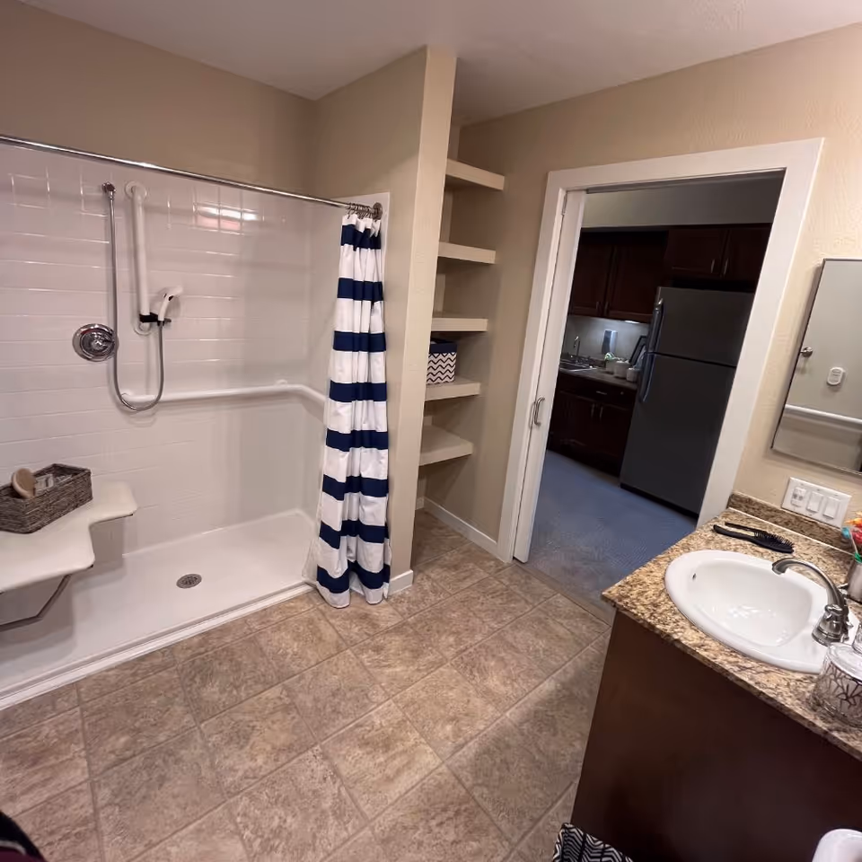 Accessible bathroom with a walk-in shower with grab bars and a blue-and-white striped curtain, sink with a granite countertop, and open shelving looking into a kitchen.