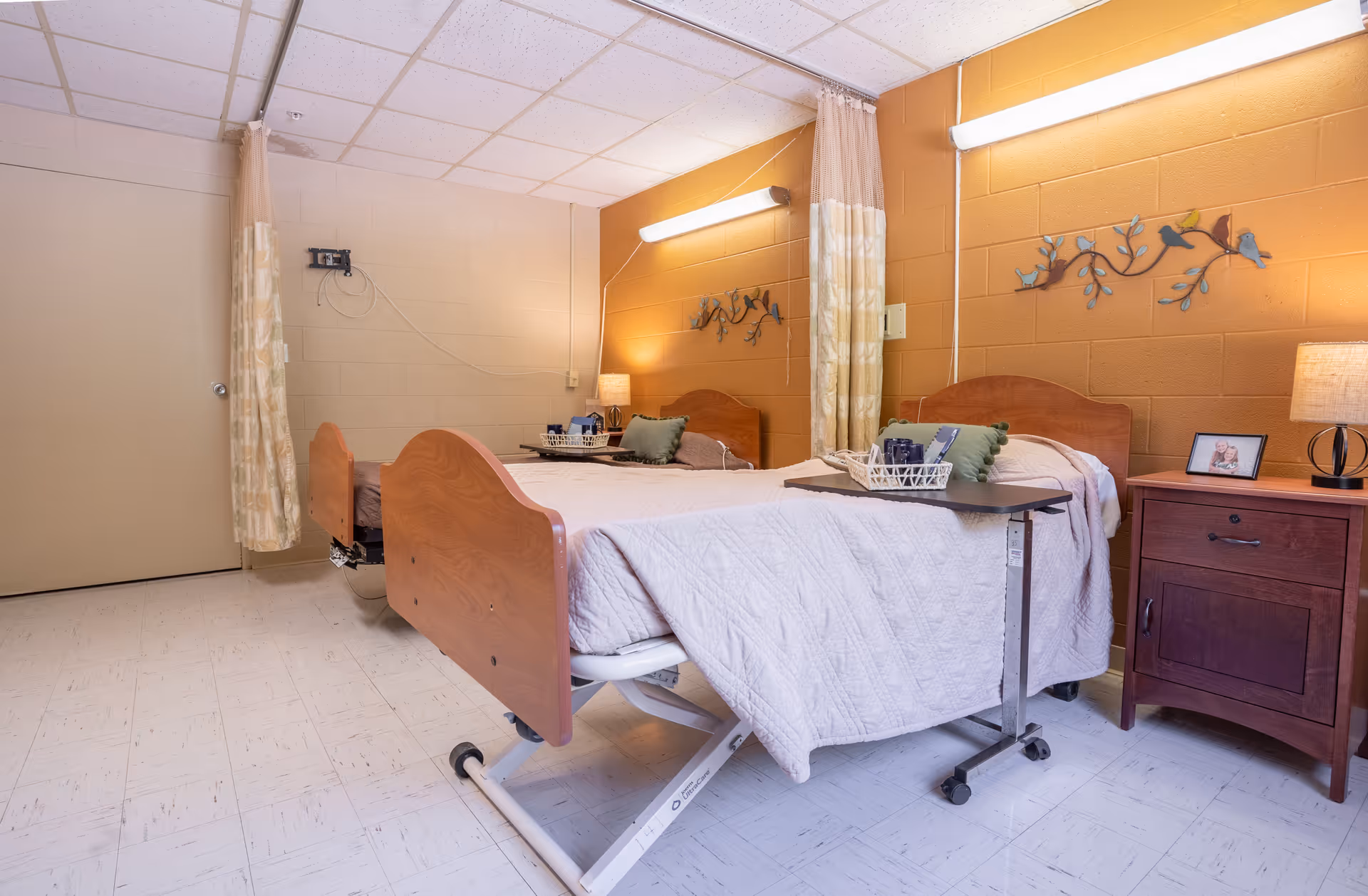 A neatly arranged senior care facility bedroom with a hospital-style bed covered in a white quilt. The bed has a green pillow and a tray table with a basket on it. There is a wooden nightstand with a lamp and a framed photo. The walls are painted in a warm yellow color with a decorative metal wall art featuring birds on branches. A curtain hangs near the door, and the floor is covered with light-colored tiles.