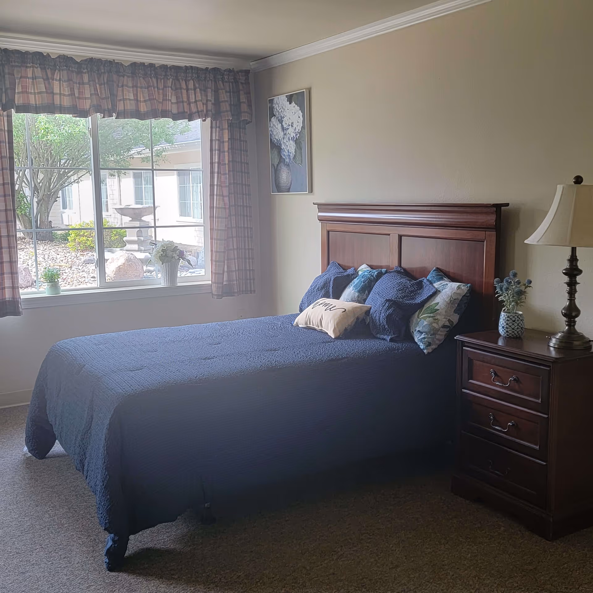 A bedroom with a wooden bed frame and a blue quilted bedspread. The bed has several decorative pillows, including one with the word 'home' on it. Next to the bed is a wooden nightstand with three drawers, a table lamp, and a small vase with flowers. A window with plaid curtains shows a view of a garden with rocks, bushes, and a birdbath outside. A framed picture of white flowers hangs on the wall above the bed.