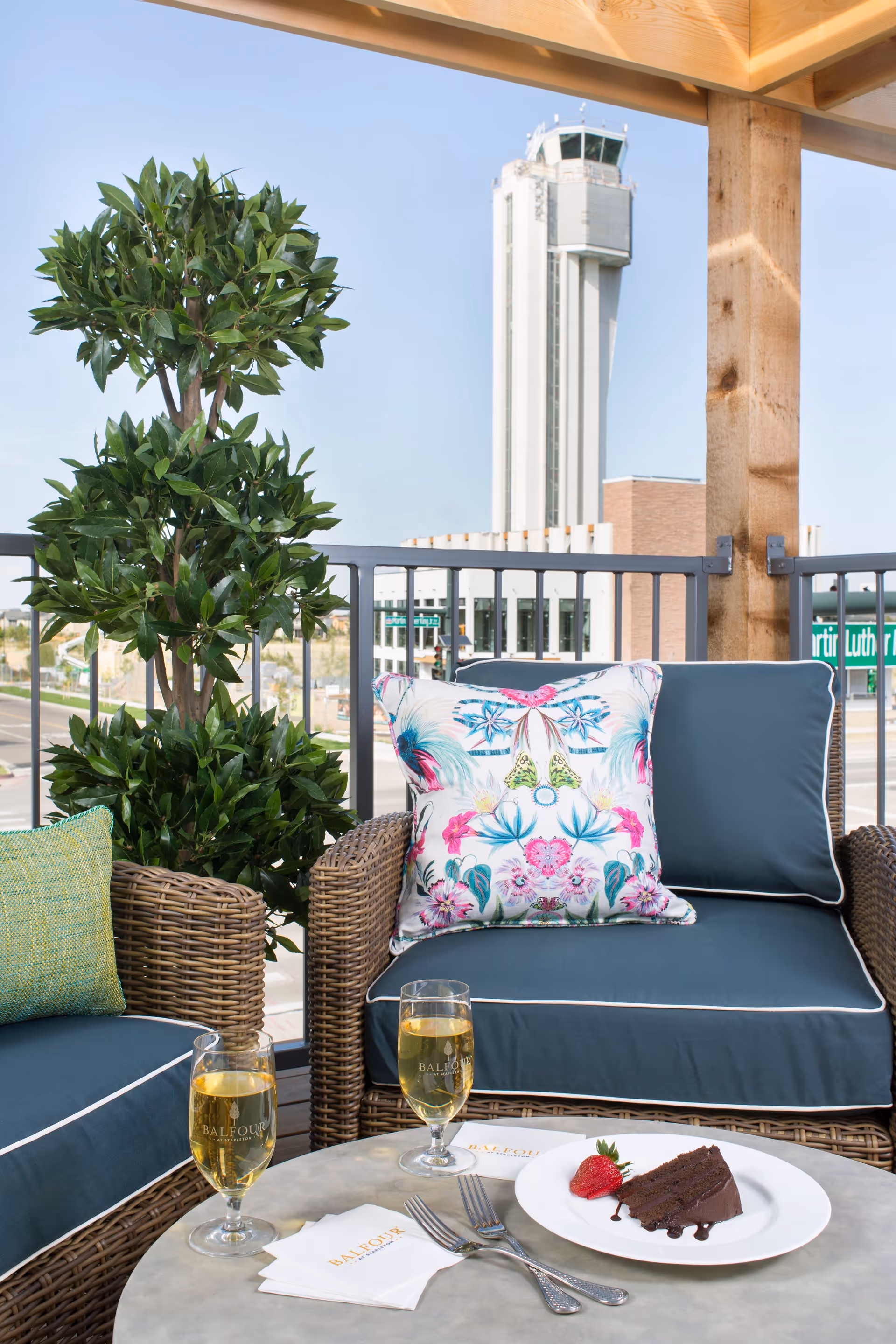 Outdoor seating area with wicker chairs featuring blue cushions and a colorful floral pillow, a small round table with two glasses of a light beverage, a plate with a slice of chocolate cake and a strawberry, and a potted green plant. In the background, there is a tall control tower and a street scene under a clear sky.