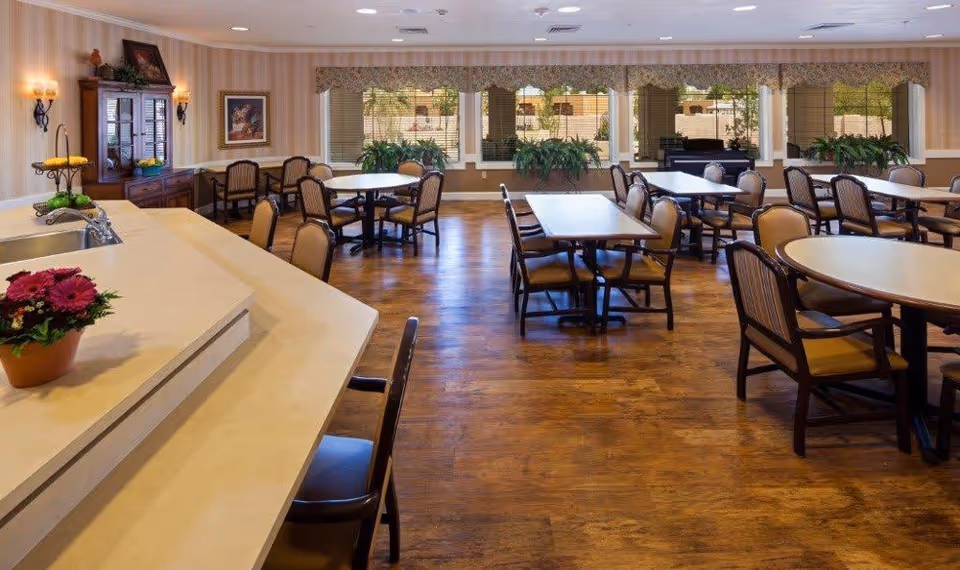 Bright dining room with round and rectangular tables and chairs, a countertop with flowers, and large windows overlooking greenery.