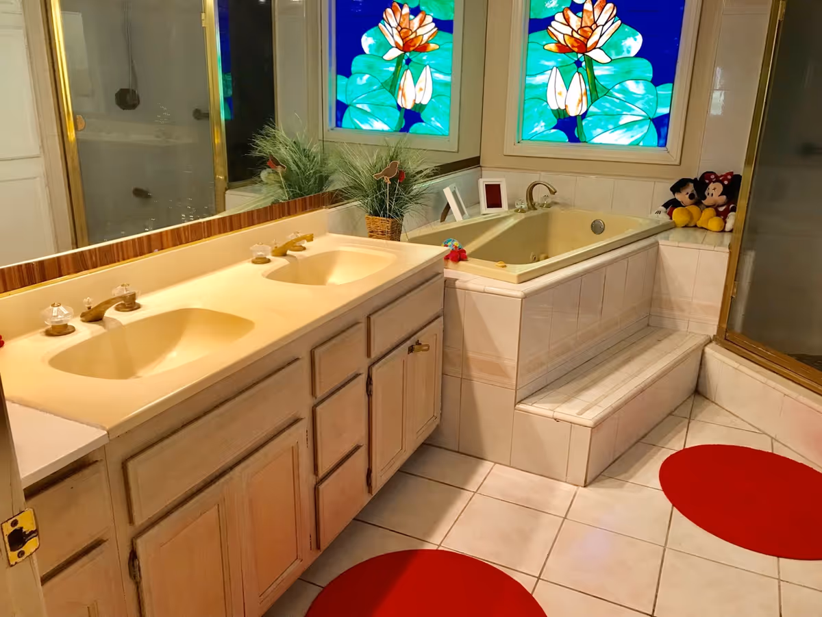 Bathroom with a double sink vanity, a built-in bathtub with steps, a glass-enclosed shower, two stained glass windows with floral designs, red circular rugs on the tiled floor, and decorative items including plants and stuffed Mickey and Minnie Mouse toys.