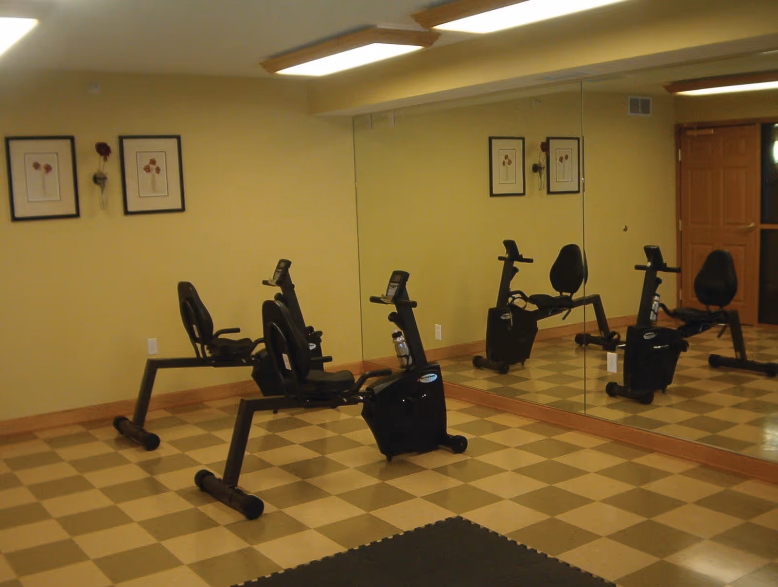 A small exercise room with two black recumbent stationary bikes on a checkered tile floor. The walls are painted yellow and decorated with two framed flower pictures and a single artificial flower in a wall-mounted vase. One wall is covered with a large mirror reflecting the bikes and pictures. There is a wooden door in the background.