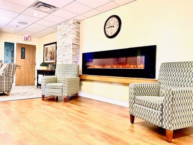A cozy lounge area with two patterned armchairs facing a modern electric fireplace mounted on a light yellow wall. Above the fireplace is a round clock. The floor is wooden, and there is a stone pillar and a wooden door with an exit sign in the background.