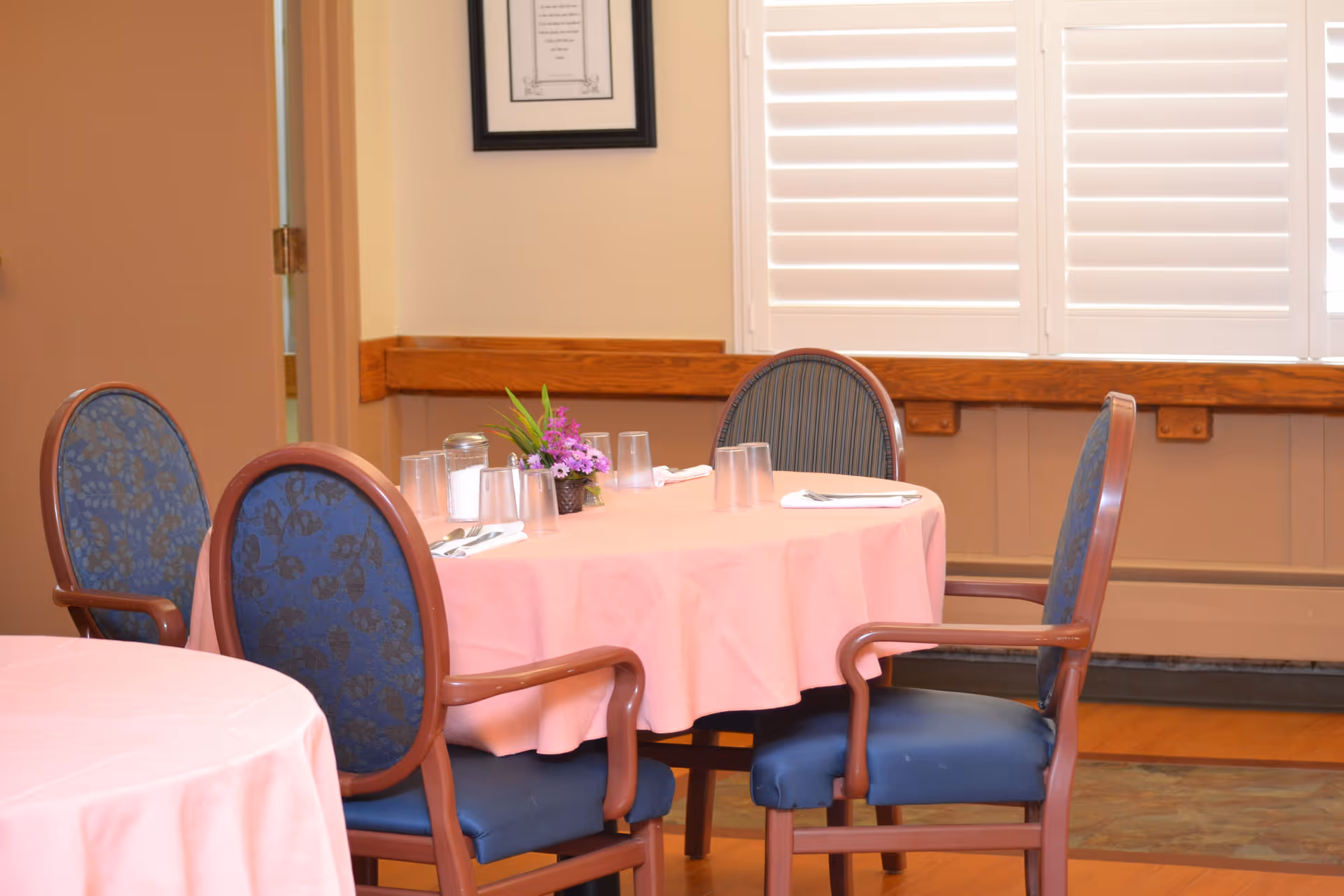 A dining area with a round table covered with a pink tablecloth, surrounded by four wooden chairs with blue cushioned seats and backs. The table is set with clear drinking glasses, silverware, napkins, and a small floral centerpiece. The background shows a window with white plantation shutters and a framed picture on the wall.