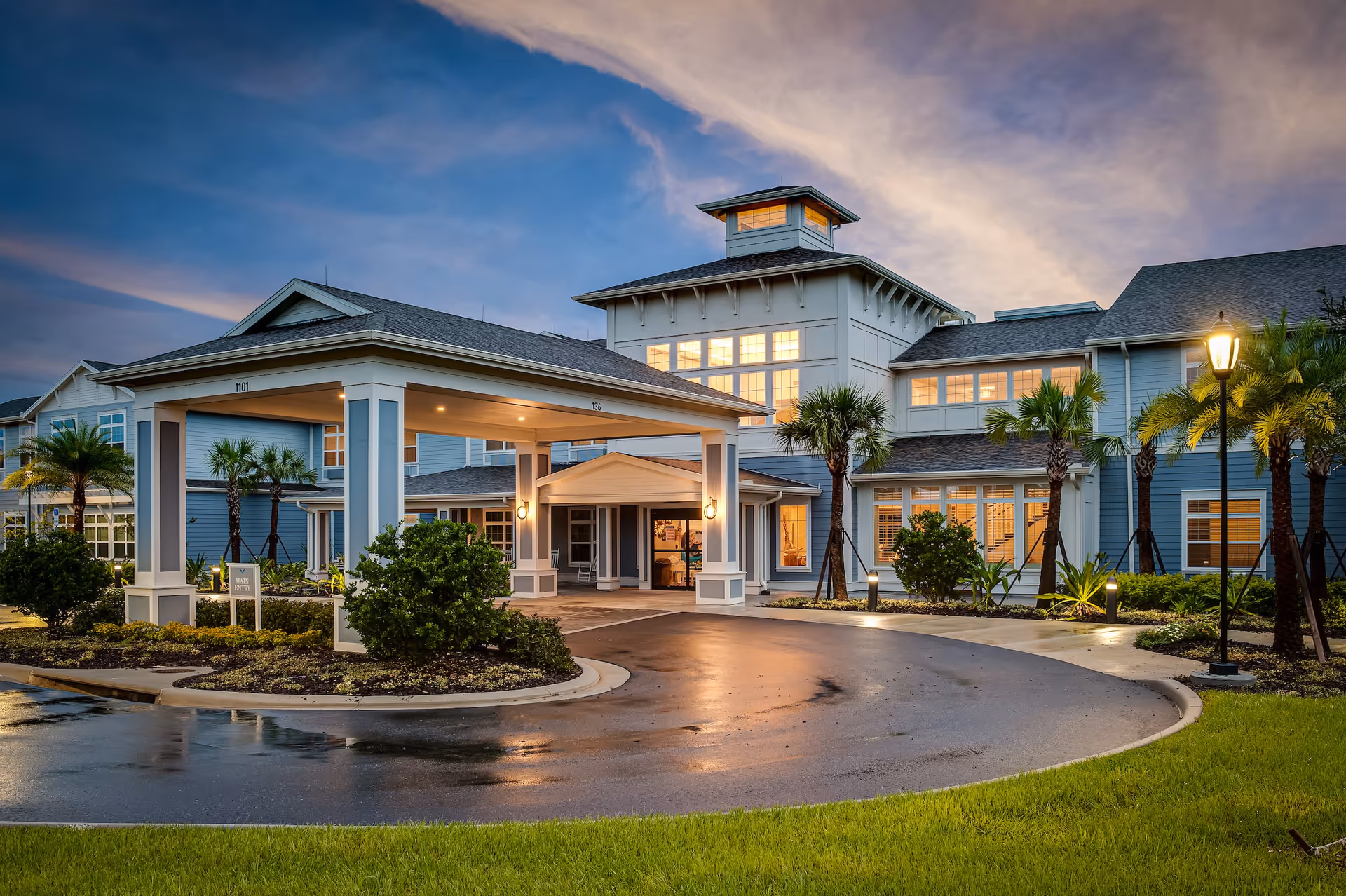 Exterior view of a senior living facility named Sancerre at Orange City during dusk, showing a large entrance with a covered drop-off area, well-lit windows, palm trees, and a curved driveway with wet pavement reflecting the lights.