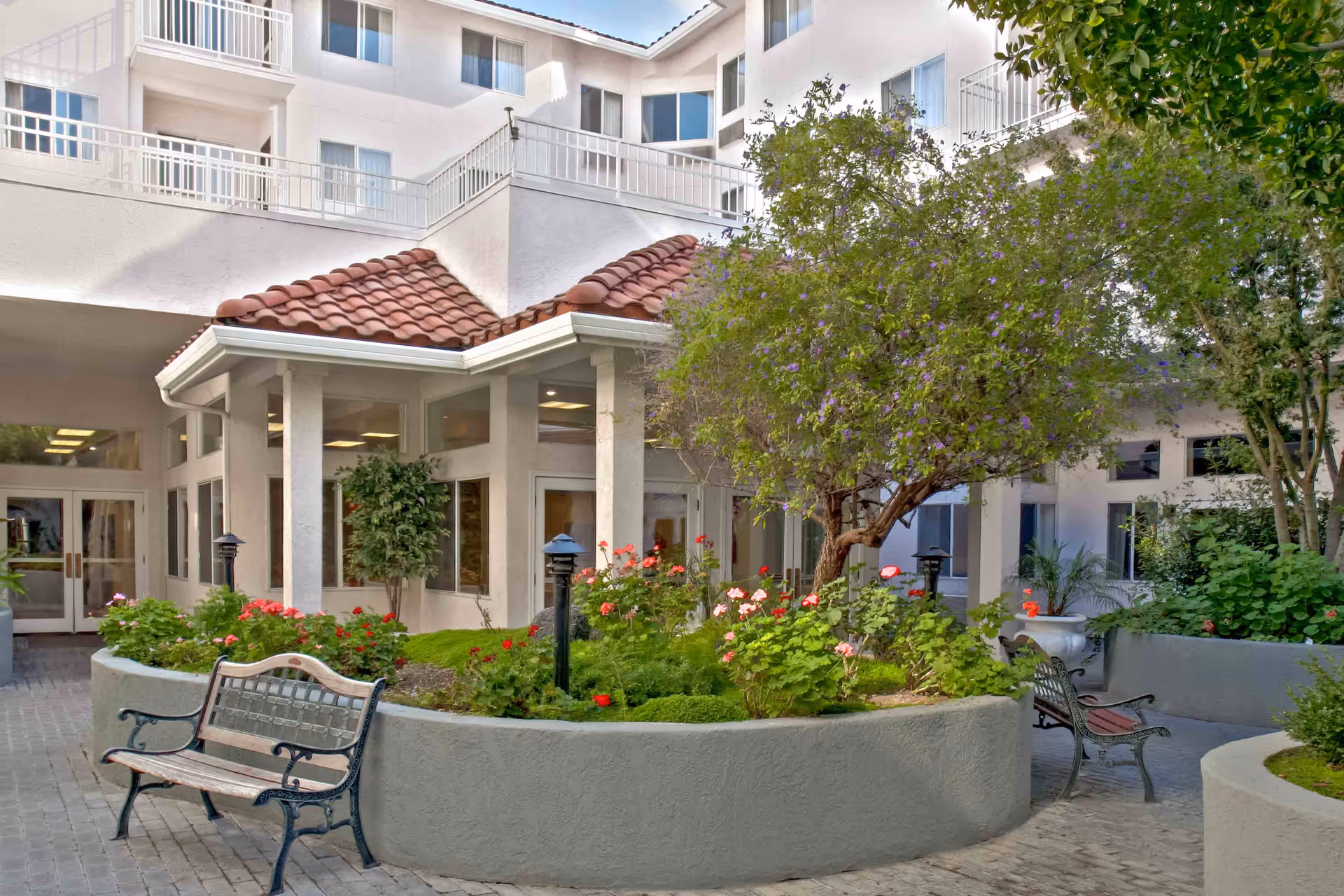 Outdoor courtyard area of Holiday Warburton Place featuring a raised garden bed with flowering plants and a tree, surrounded by benches and a paved walkway. The building exterior with white walls and red-tiled roof is visible in the background.