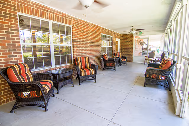 A covered outdoor patio area with six black wicker chairs featuring red and orange striped cushions. There are two small glass-top wicker tables between some of the chairs. The patio has a brick wall with windows on one side and large screened windows on the other side. Ceiling fans are mounted on the white ceiling.