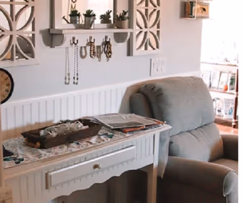 A cozy sitting area with a gray recliner beside a white console table holding magazines and a decorative tray under a wall shelf with hooks and small plants.