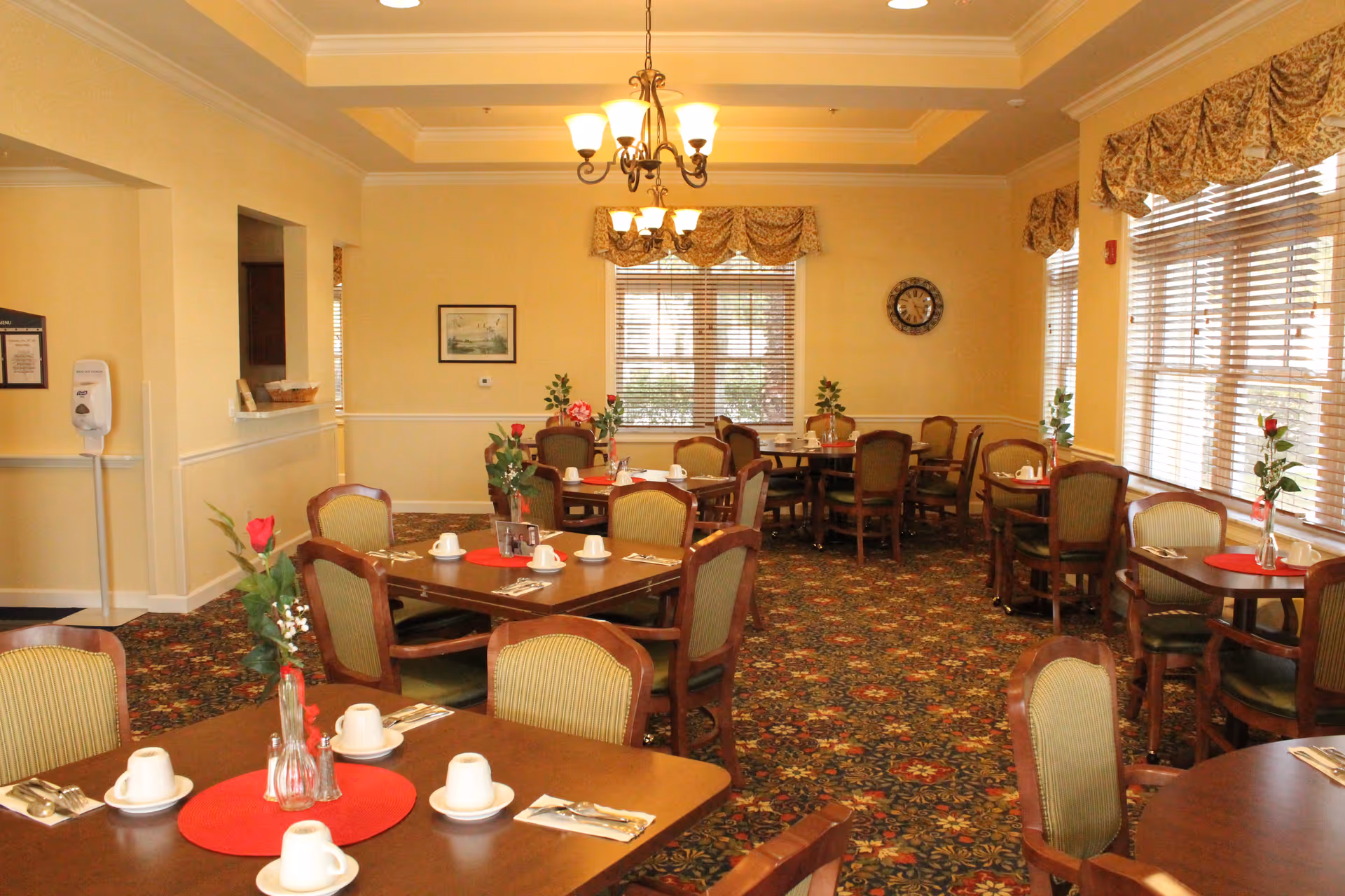 A dining room in a senior living facility with multiple wooden tables and chairs. Each table is set with white cups, saucers, silverware, and red placemats. There are vases with red roses on the tables. The room has beige walls, large windows with blinds and patterned valances, a patterned carpet, and a chandelier hanging from the ceiling.