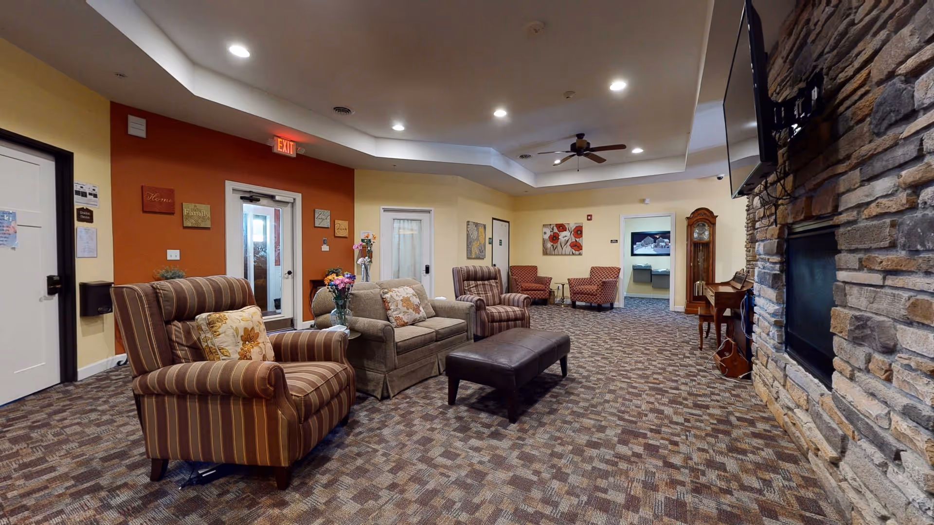 A spacious and cozy living room area in a retirement facility with patterned carpet flooring, several upholstered armchairs and a sofa with floral cushions, a dark ottoman in the center, a stone fireplace with a mounted TV above it, a wooden grandfather clock, and a piano. The walls are painted in warm tones with decorative wall art and an exit door visible.