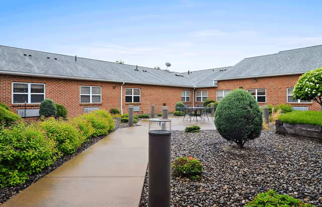 Outdoor courtyard area of a senior living facility with a paved walkway, landscaped bushes, small trees, and patio tables with chairs. The building is a single-story brick structure with multiple windows and a gray shingled roof under a clear blue sky.