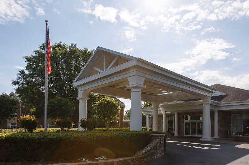 Entrance of a senior living facility with a covered drop-off area supported by large white columns, an American flag on a flagpole, and well-maintained landscaping including bushes and trees under a partly cloudy sky.