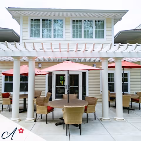 Outdoor patio area at All American Assisted Living At Enfield featuring round tables with wicker chairs and red umbrellas under a white pergola attached to a two-story building with large windows.