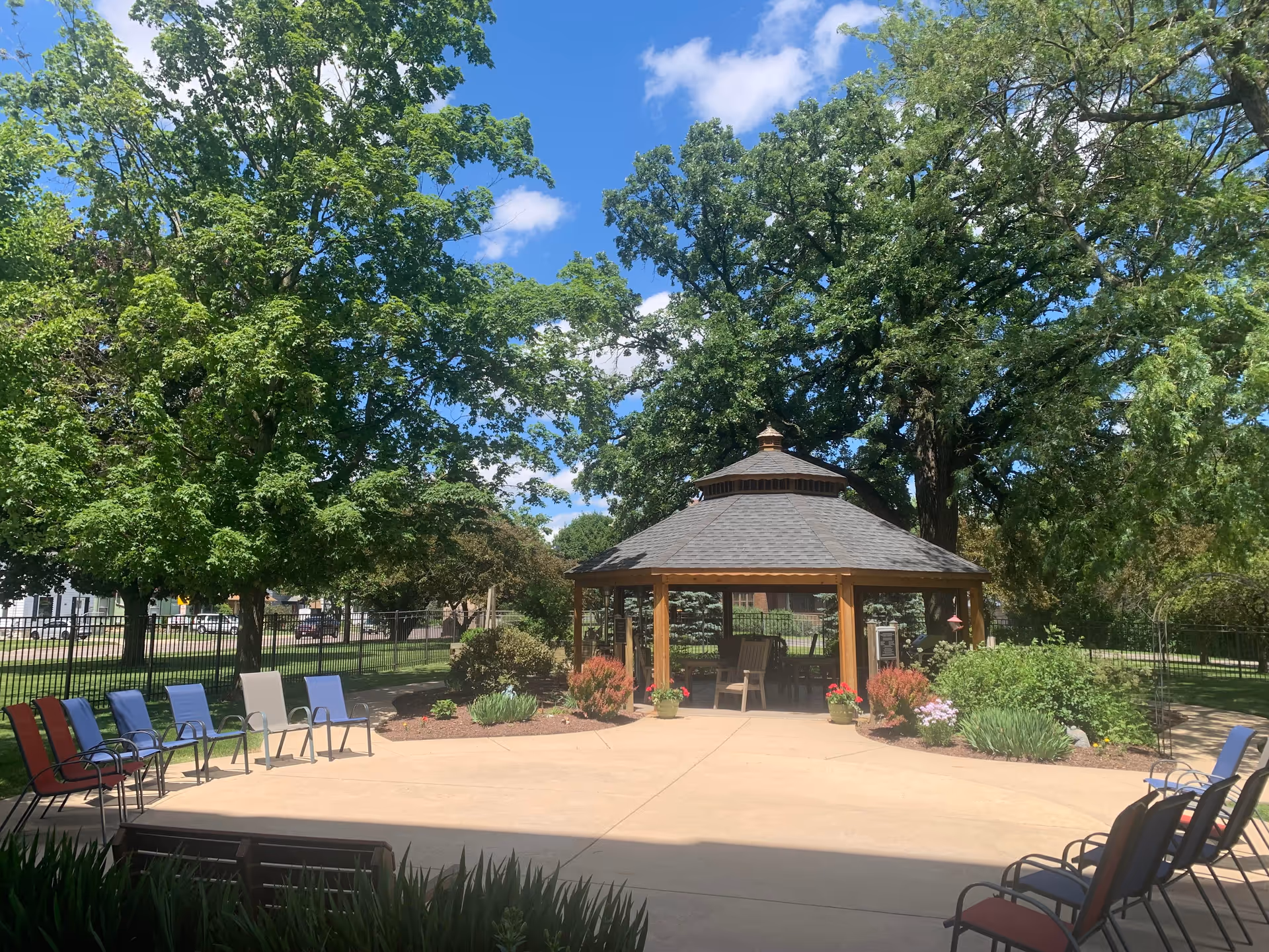 Outdoor patio area with a wooden gazebo surrounded by trees and plants. Several chairs are arranged in a semicircle on the concrete patio under a clear blue sky.