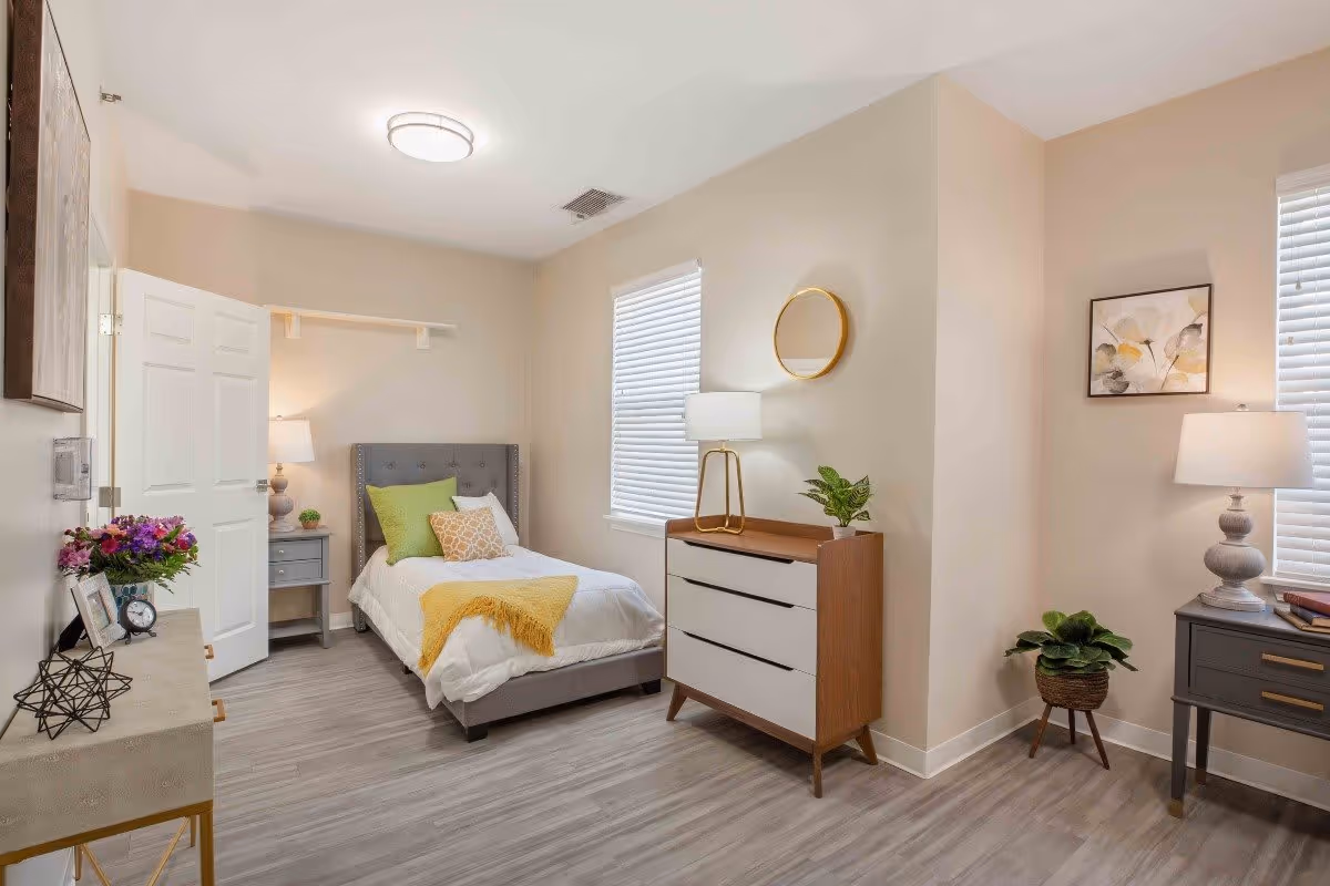 A bright and neatly arranged bedroom with a single bed featuring white bedding, green and patterned pillows, and a yellow throw blanket. The room includes a gray nightstand with a lamp and small plant, a wooden dresser with a lamp and round mirror above it, another gray side table with a lamp and books, a potted plant on the floor, and light-colored walls and flooring.